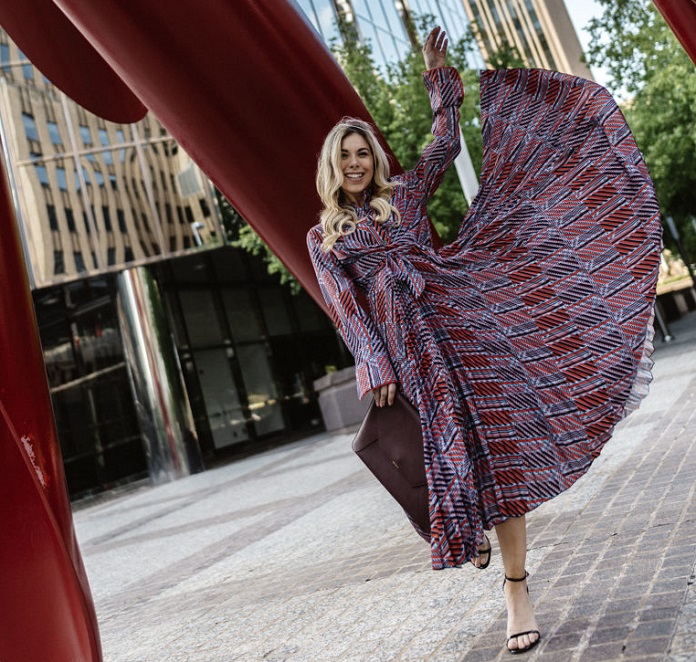 picture of woman standing under an outside art scolpture near a building waving her printed dress 